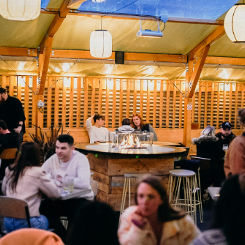 Cozy evening dining experience at Leaven Pizza in Hereford, featuring groups of people enjoying drinks and wood-fired pizza in a warm, stylish outdoor seating area with hanging lanterns.